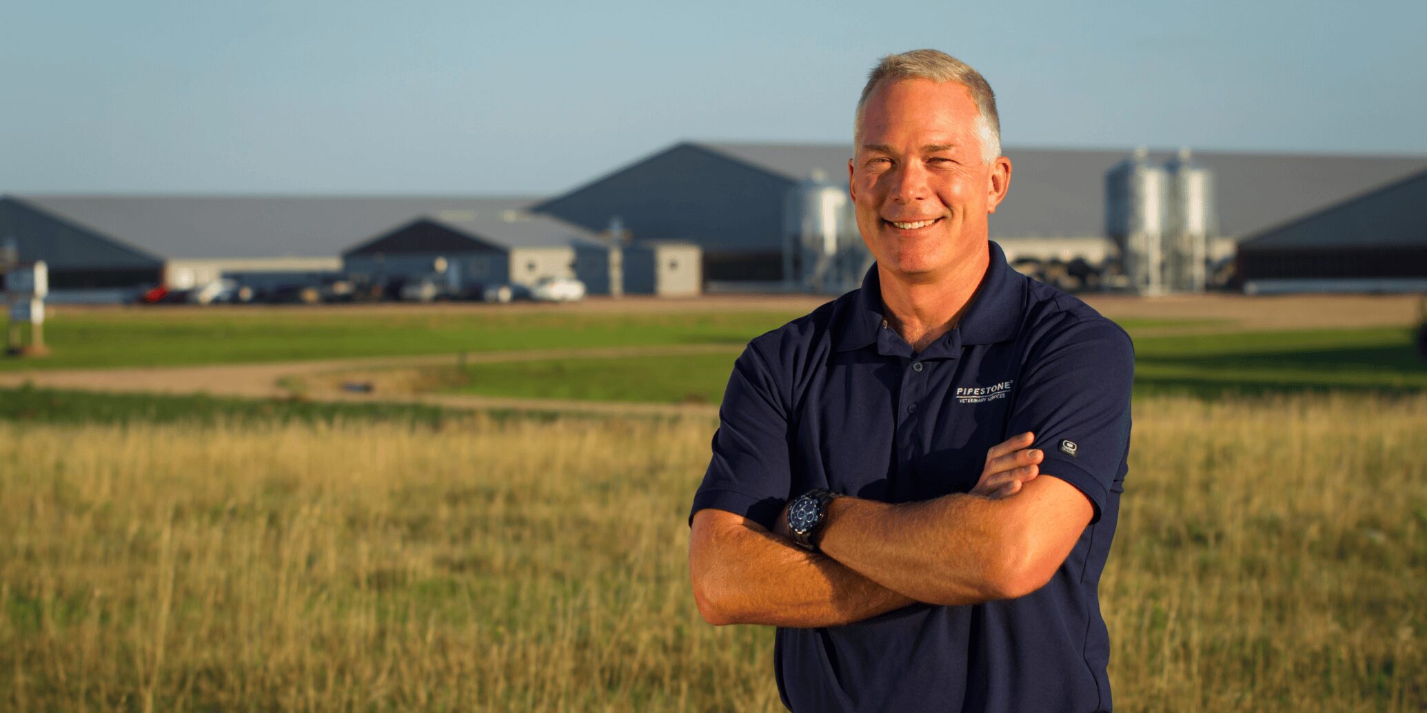 A man stands in a grassy area with swine barns behind him in the distance, smiling with his arms crossed.