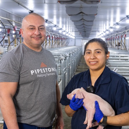Two barn workers stand in a barn aisle with one holding a piglet.