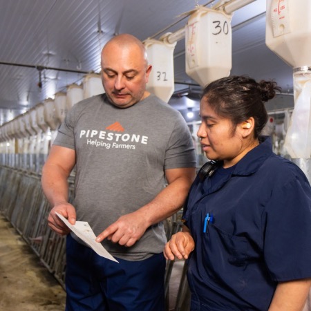A barn manager has a conversation and points at swine data he is holding as he talks to a female farm worker in a swine barn aisle.
