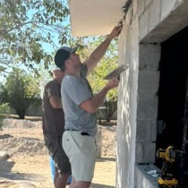 Two pipestone team members stand in front of a brick wall and spread cement over it.