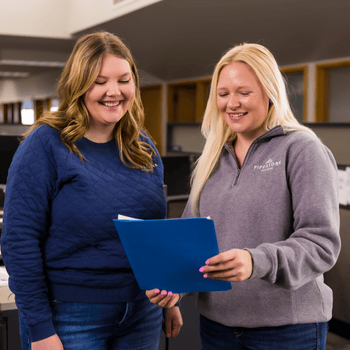 Two pipestone employees in the office stand and look at scholarship papers in front of them.