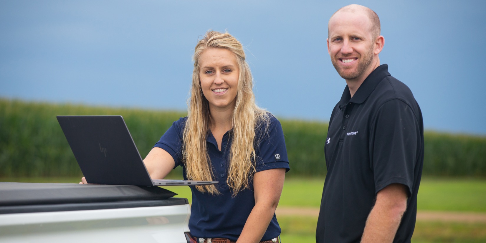 Two employees smiling outside standing by truck bed and laptop.