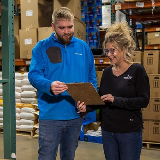 Two animal health warehouse workers stand next to product and look at order list.