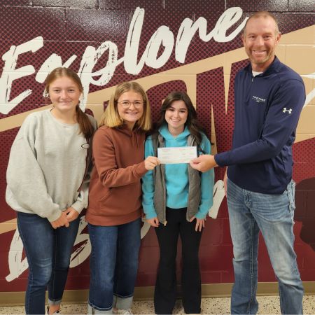 A pipestone employee visits a local school and gives a check to three students to go toward their Fuel4Kids backpack program.