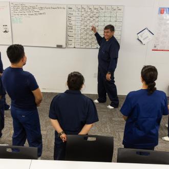 A barn manager stands in the front of the room to address a group of workers while pointing at a whiteboard.