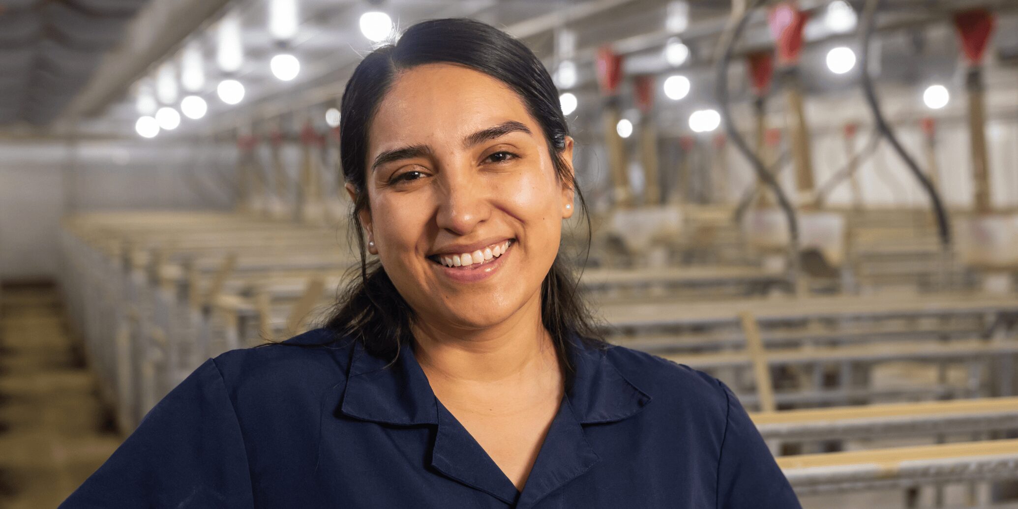 A swine barn worker smiling with a farrowing room behind her.