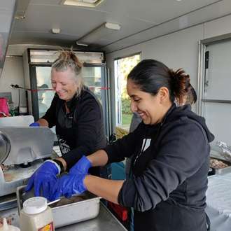 Two pipestone team members stand in a trailer and cook pork products to sell at an event.