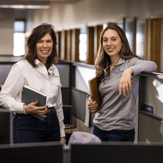 Two pipestone office workers stand by a desk smiling.