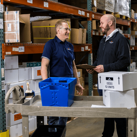 Two warehouse workers stand in front of a cart with boxes of animal health products.