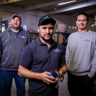 Three team members standing in a repair shop with one holding a mechanical part.
