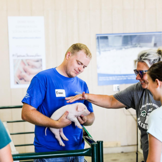 A volunteer holding a piglet while a fairgoer touches it.