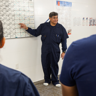 A swine barn manager stands in front of the room pointing at a calendar while the farm workers listen.
