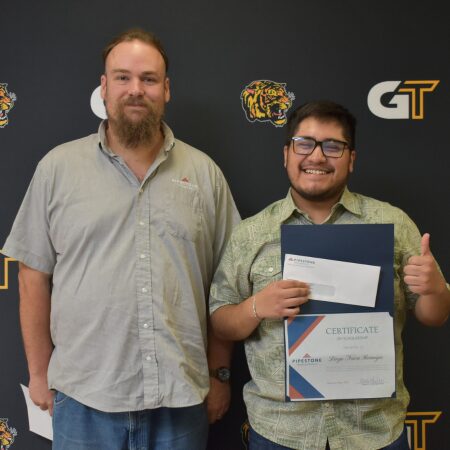 Two men stand in front of a backdrop as one holds a scholarship and gives a thumbs up.