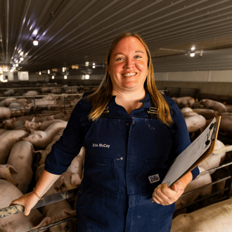 A veterinary physician assistant stands with a clipboard in front of a group of pigs in a barn.