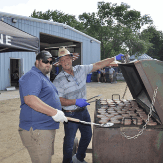 Two men operate a grill outside cooking burgers for a company picnic.