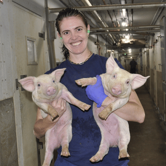 A pig veterinarian is holding two piglets in her arms inside a swine facility.